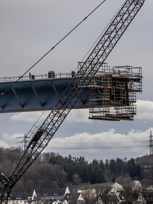 Untersicht auf zwei Brückenelemente, die per Kran zusammengeführt werden. Am unteren Bildrand ist eine Ortschaft zu sehen. Untersicht auf zwei Brückenelemente, die per Kran zusammengeführt werden. Am unteren Bildrand ist eine Ortschaft zu sehen.