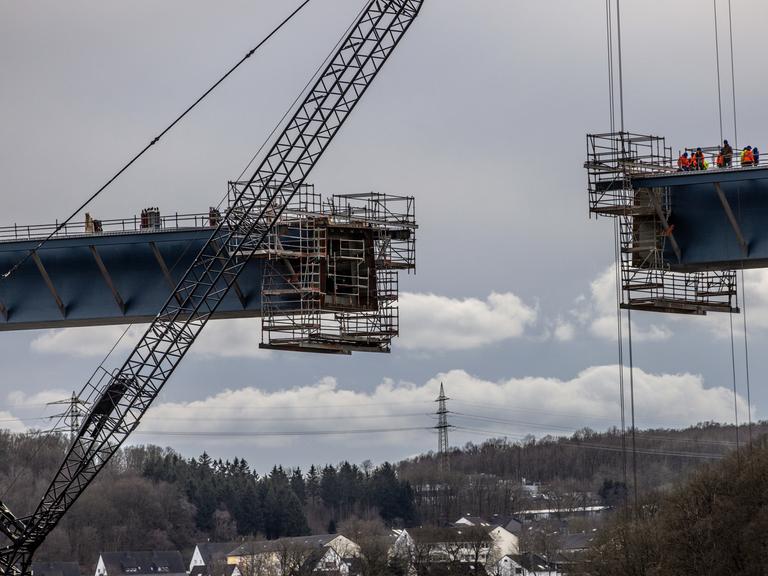 Untersicht auf zwei Brückenelemente, die per Kran zusammengeführt werden. Am unteren Bildrand ist eine Ortschaft zu sehen. Untersicht auf zwei Brückenelemente, die per Kran zusammengeführt werden. Am unteren Bildrand ist eine Ortschaft zu sehen.