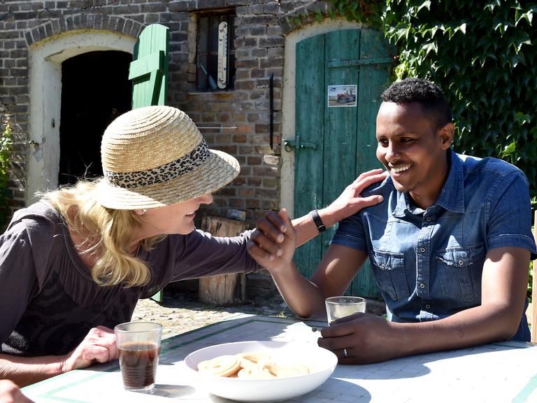 Eine Frau mit Hut, Hildegard Nies-Nachtsheim aus Brandenburg, sitzt mit einem Mann, Jarmal aus Somalia, an einem Tisch in einem Garten, isst und trinkt. Sie legt ihm die Hand auf die Schulter und beide lächeln.