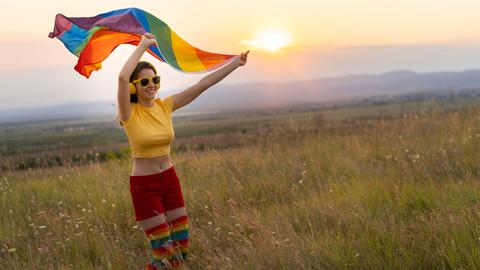 Glückliche junge Frau in sommerlicher bunter Kleidung mit Kopfhörern und Regenbogenfahne vor einem Sonnenuntergang.