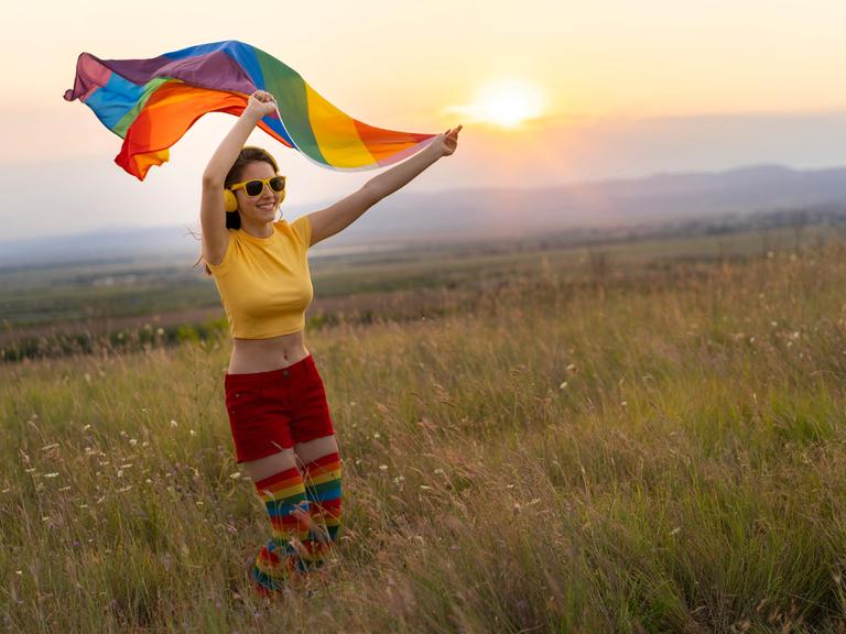 Glückliche junge Frau in sommerlicher bunter Kleidung mit Kopfhörern und Regenbogenfahne vor einem Sonnenuntergang.