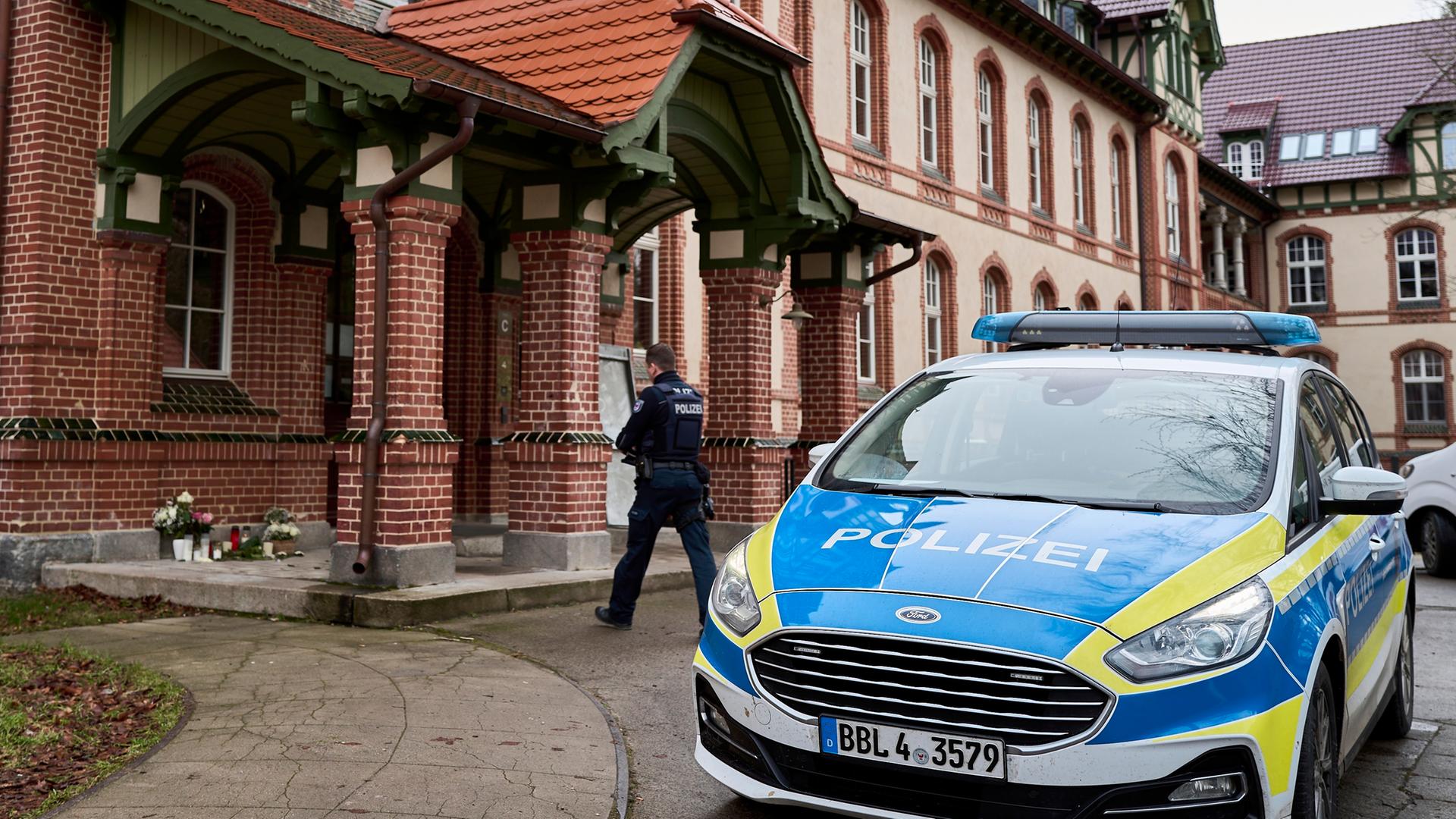 Beelitz: Blumen und Kerzen stehen vor dem Eingang eines Hauses nach einem Gewaltverbrechen, während ein Polizist am Tatort eintrifft. 