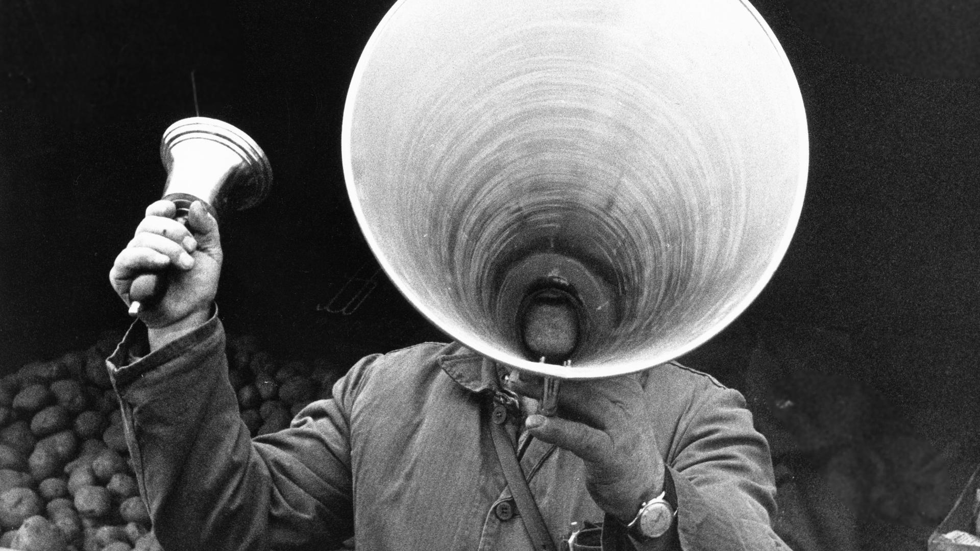 Ein Marktschreier hebt ein riesiges Megaphon vor seinen Mund, in der rechten Hand hält er eine Glocke. 