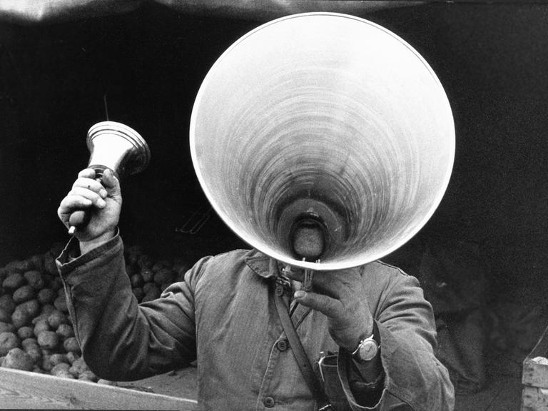 Ein Marktschreier hebt ein riesiges Megaphon vor seinen Mund, in der rechten Hand hält er eine Glocke. 