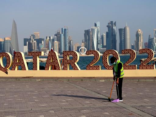 Ein Arbeiter räumt vor einem "Qatar 2022"-Schild Müll weg. Im Hintergrund die Skyline von Doha.