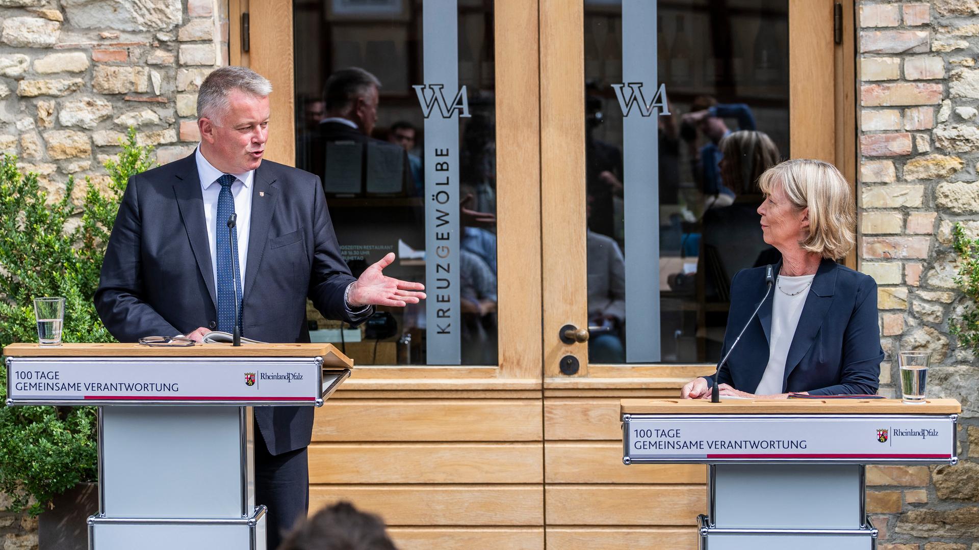 Die beiden stehen am Eingang eines Steinhauses hinter Rednerpulten. Schnieder sagt etwas, gestikuliert und wendet sich Ahnen zu. Die beiden stehen am Eingang eines Steinhauses hinter Rednerpulten. Schnieder sagt etwas, gestikuliert und wendet sich Ahnen zu.