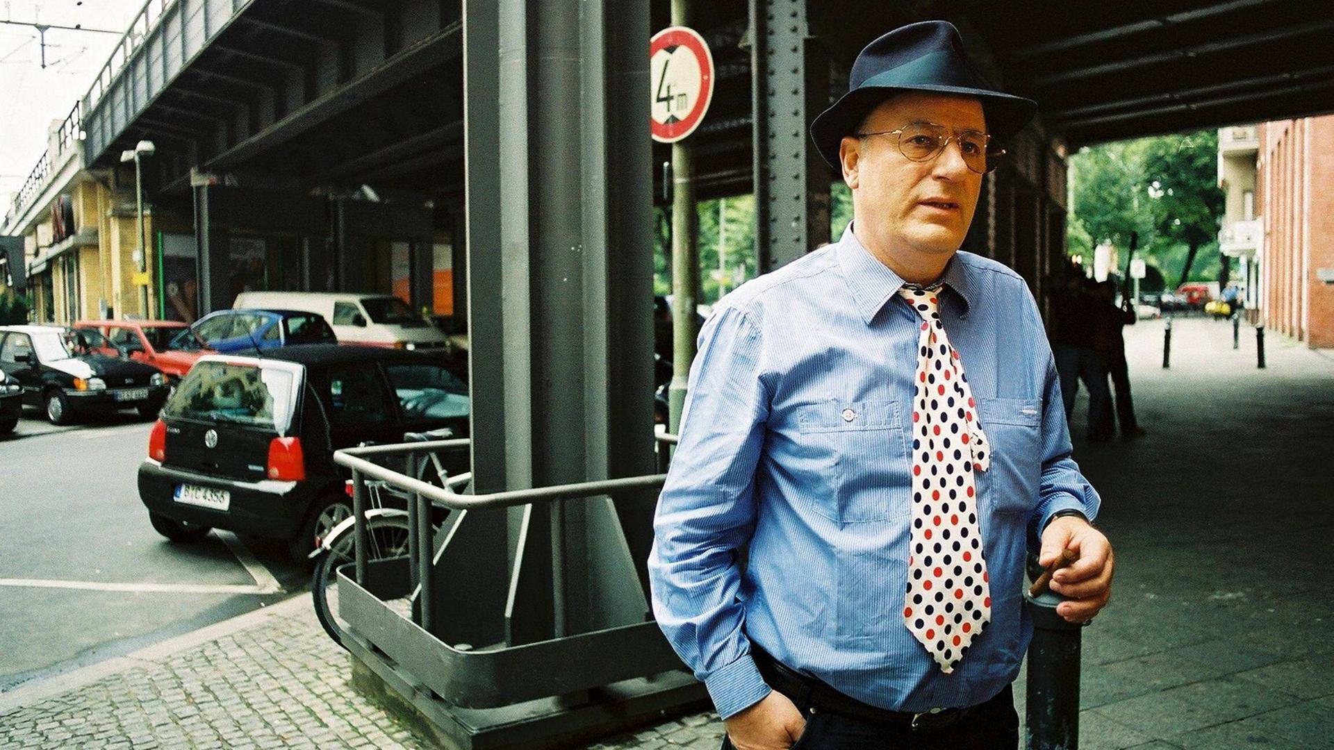 Manfred Krug bei einem Pressetermin am Savignyplatz in Berlin. Er trägt ein blaues Hemd, dazu eine weisse Krawatte mit schwarzen und roten Punkten.