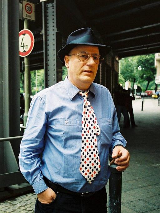 Manfred Krug bei einem Pressetermin am Savignyplatz in Berlin. Er trägt ein blaues Hemd, dazu eine weisse Krawatte mit schwarzen und roten Punkten.