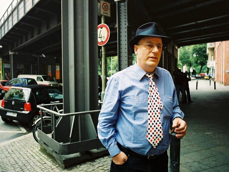 Manfred Krug bei einem Pressetermin am Savignyplatz in Berlin. Er trägt ein blaues Hemd, dazu eine weisse Krawatte mit schwarzen und roten Punkten.