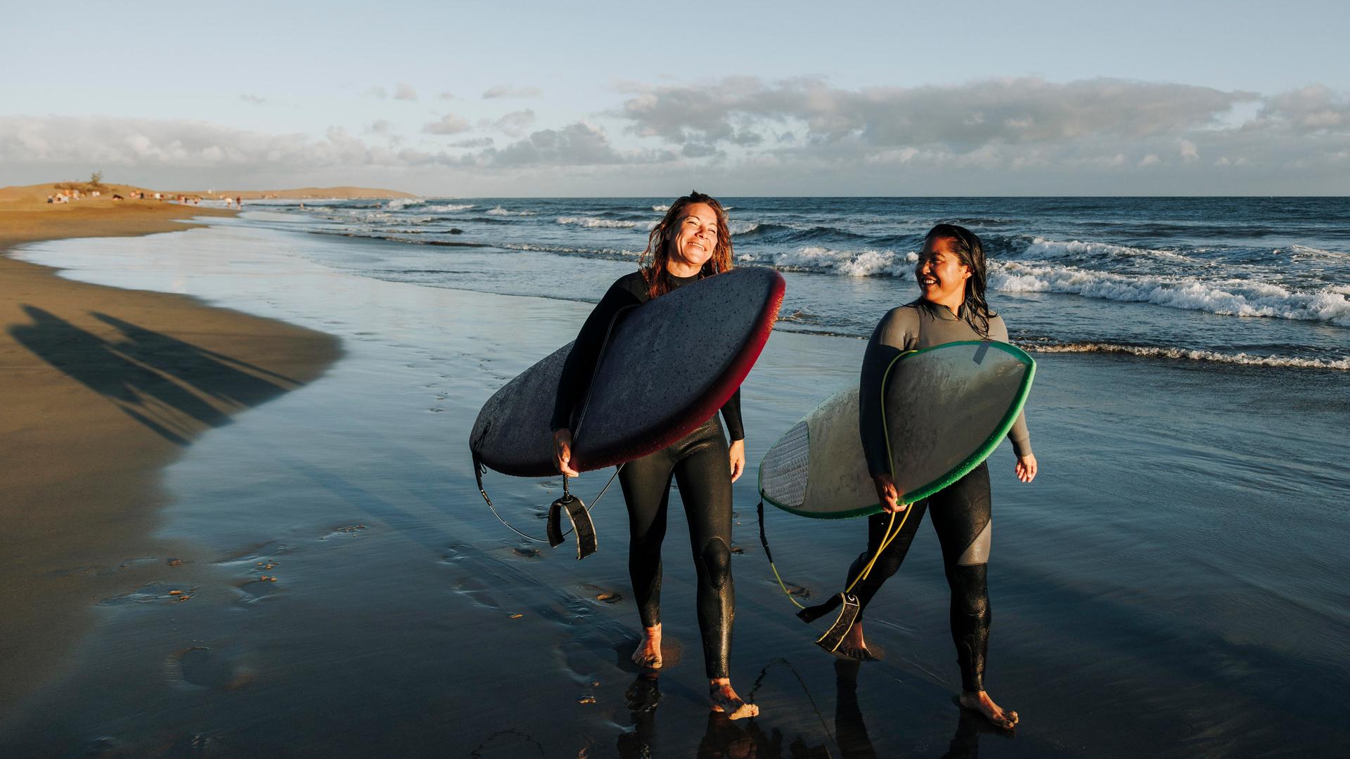 Zwei Frauen laufen mit ihren Surfboards nebeneinander an einem Strand entlang.