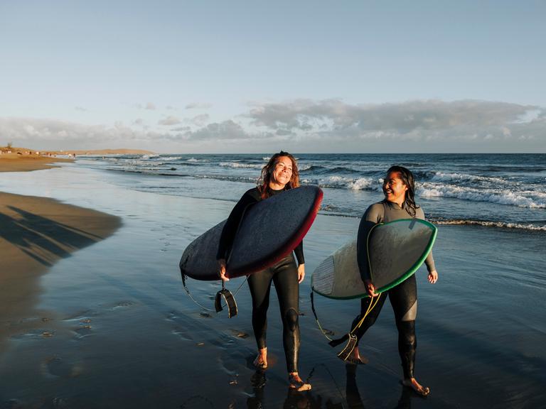 Zwei Frauen laufen mit ihren Surfboards nebeneinander an einem Strand entlang.