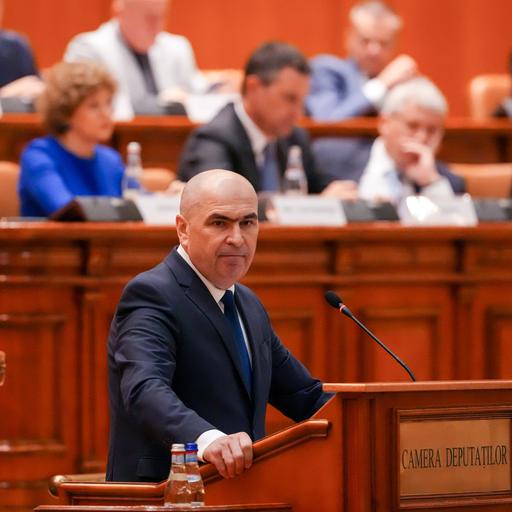 Der rumänische Premierminister Ilie Bolojan spricht in Bukarest vor einer Parlamentssitzung. Hinter ihm sitzen mehrere Personen. 