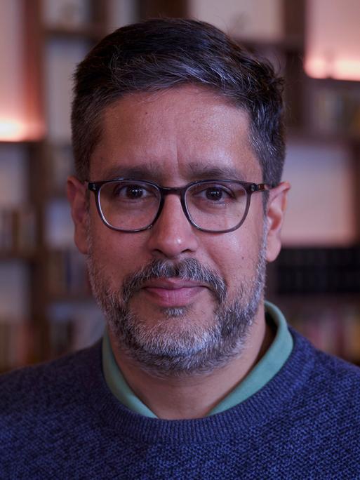 Der deutsche Journalist und Autor Hasnain Niels Kazim blickt in die Kamera. Im Bildhintergrund ist unscharf eine Bücherwand zu erkennen Der deutsche Journalist und Autor Hasnain Niels Kazim blickt in die Kamera. Im Bildhintergrund ist unscharf eine Bücherwand zu erkennen