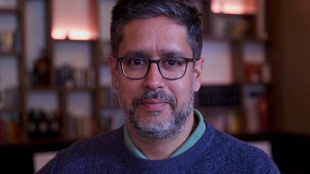 Der deutsche Journalist und Autor Hasnain Niels Kazim blickt in die Kamera. Im Bildhintergrund ist unscharf eine Bücherwand zu erkennen