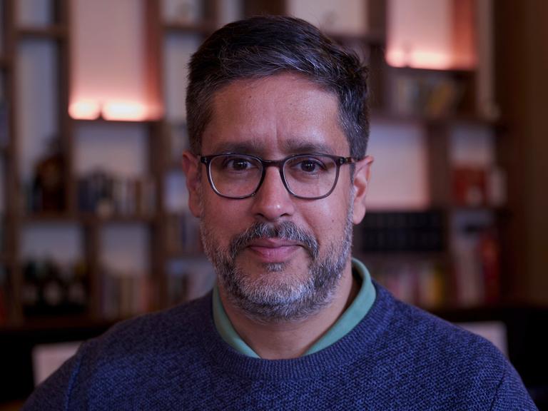 Der deutsche Journalist und Autor Hasnain Niels Kazim blickt in die Kamera. Im Bildhintergrund ist unscharf eine Bücherwand zu erkennen Der deutsche Journalist und Autor Hasnain Niels Kazim blickt in die Kamera. Im Bildhintergrund ist unscharf eine Bücherwand zu erkennen