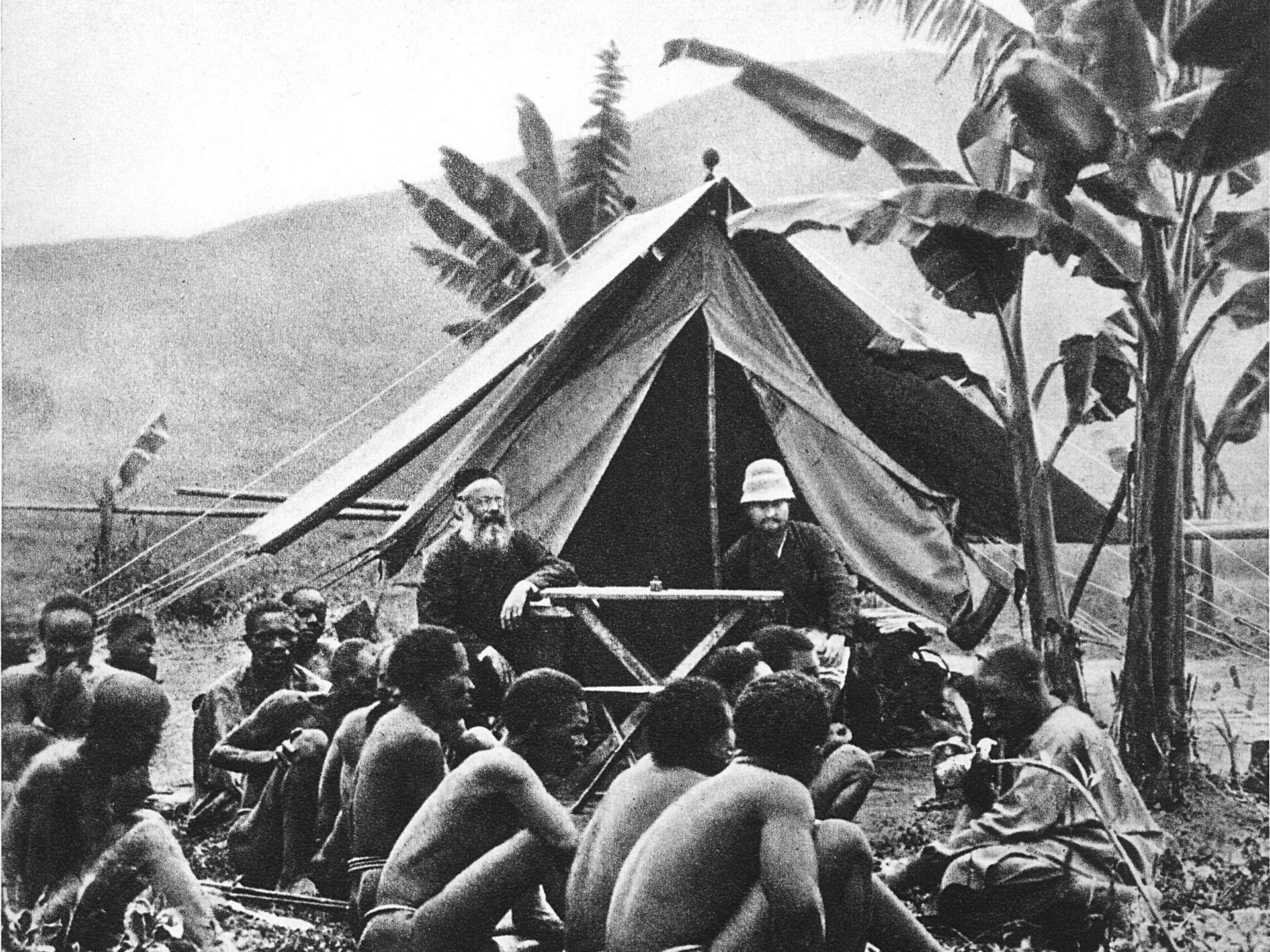 Historisches Schwarzweißfoto: Deutsche Missionare sitzen in einem Zelt. Davor sitzen Männer der lokalen Bevölkerung auf dem Boden.