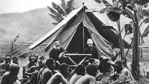 Historisches Schwarzweißfoto: Deutsche Missionare sitzen in einem Zelt. Davor sitzen Männer der lokalen Bevölkerung auf dem Boden.