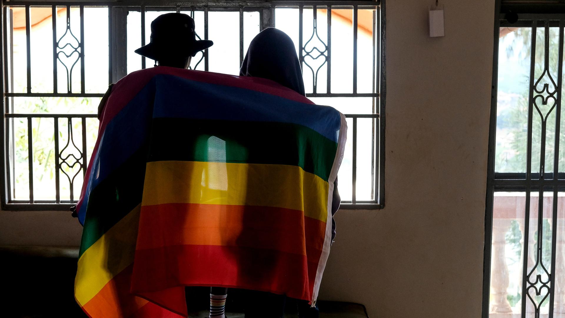 Ein Paar sitzt mit dem Rücken zur Kamera in einem Gebäude vor einem Fenster. Beide haben sich eine Pride-Flagge um die Schultern gelegt.