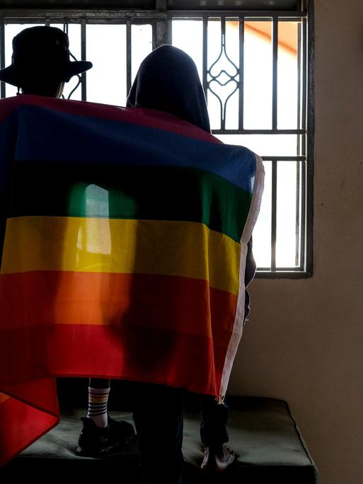 Ein Paar sitzt mit dem Rücken zur Kamera in einem Gebäude vor einem Fenster. Beide haben sich eine Pride-Flagge um die Schultern gelegt.