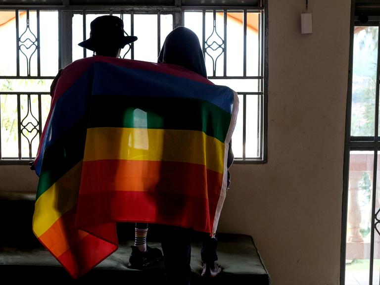 Ein Paar sitzt mit dem Rücken zur Kamera in einem Gebäude vor einem Fenster. Beide haben sich eine Pride-Flagge um die Schultern gelegt.