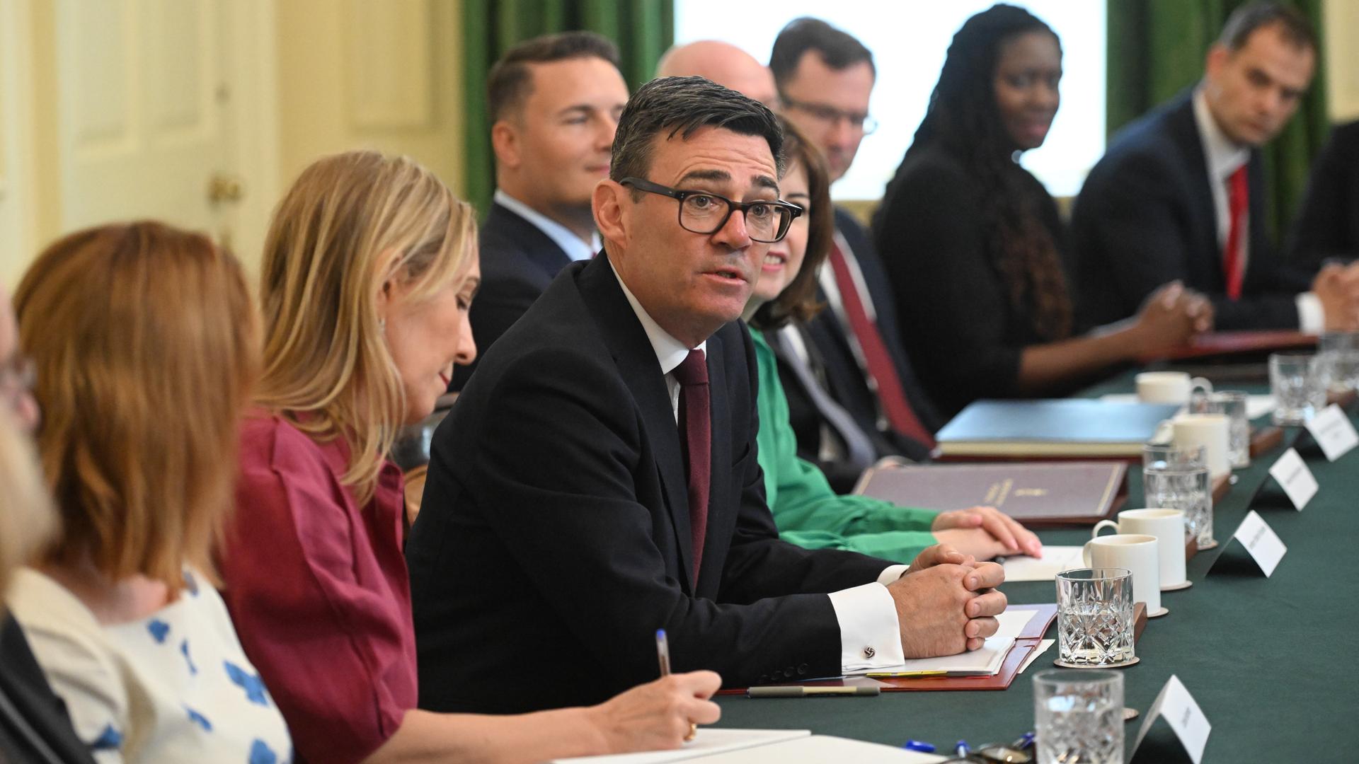 Der britische Premierminister Andy Burnham sitzt inmitten seiner Ministerinnen und Minister an einem großen Tisch. Der britische Premierminister Andy Burnham sitzt inmitten seiner Ministerinnen und Minister an einem großen Tisch.