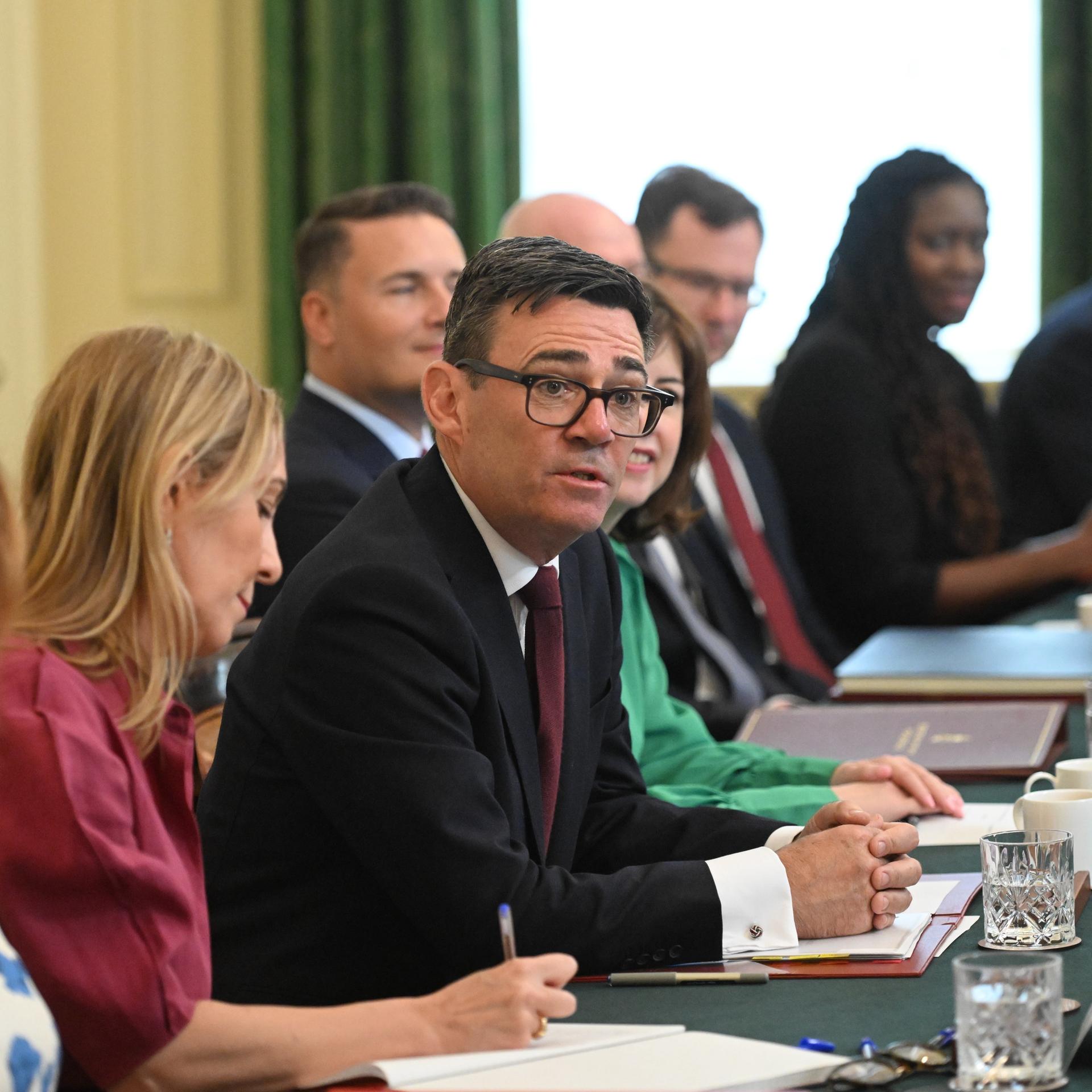 Der britische Premierminister Andy Burnham sitzt inmitten seiner Ministerinnen und Minister an einem großen Tisch.