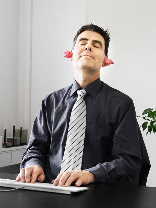 Ein Geschäftsmann sitzt grinsend am Schreibtisch. In seinen Ohren stecken selbstgebastelte Stöpsel aus Papier.
