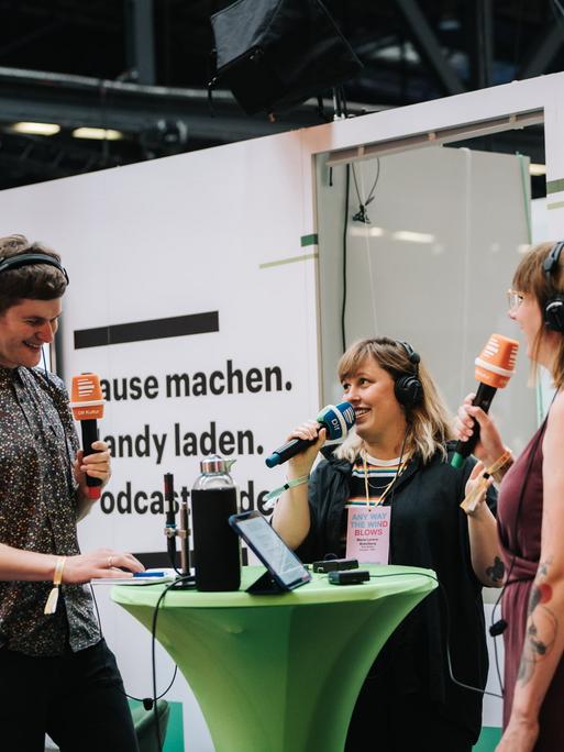 Auf einer Bühne in einer Messehalle stehen Mike Herbstreuth, Maria Lorenz-Bokelberg und Carina Schroeder in gelöster Gesprächshaltung.