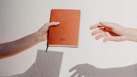 Vor einem weißen Bildhintergrund wird ein in braunes Leder gebundenes Tagebuch von einer Hand in die Nächste weitergereicht.