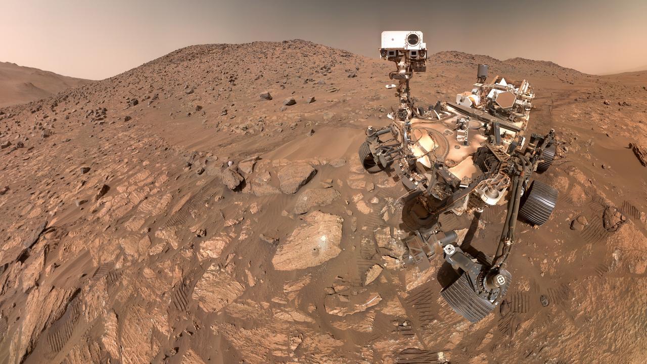 Der Rover steht auf einer steinigen Landschaft. Seine Räder haben im roten Sand Spuren hinterlassen.