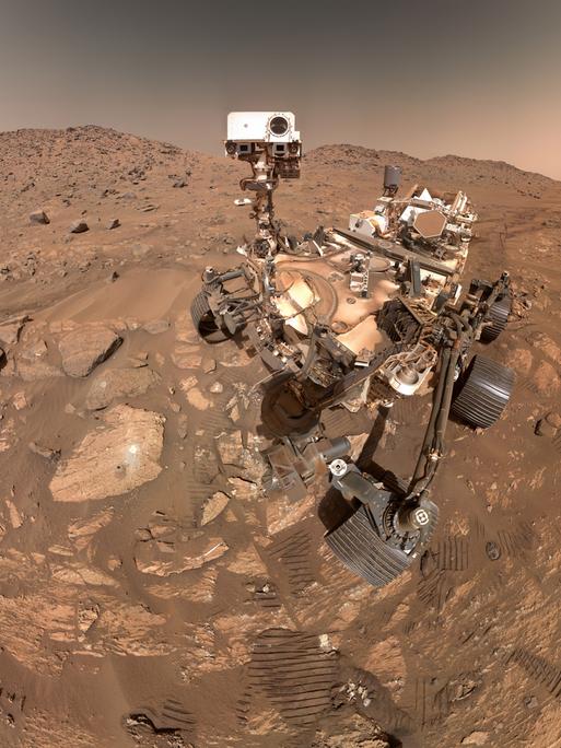 Ein Foto zeigt eine steinige rote Landschaft auf dem Mars, rechts sieht man den Forschungsrover Perseverance und Abrücke von dessen Rädern im Sand.