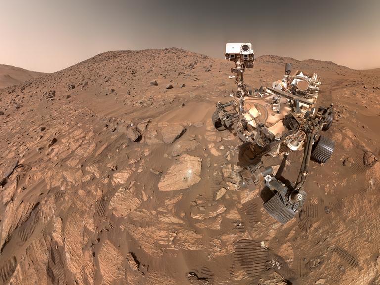 Ein Foto zeigt eine steinige rote Landschaft auf dem Mars, rechts sieht man den Forschungsrover Perseverance und Abrücke von dessen Rädern im Sand.