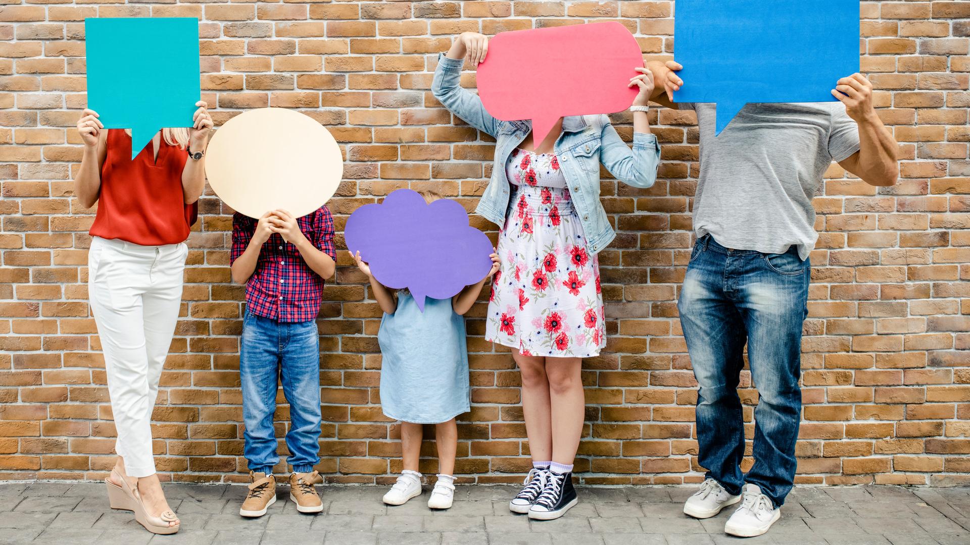 Jede Person der abgebildeten fünfköpfigen Familie hält eine Sprechblase aus farbiger Pappe vor dem Gesicht. Die Gesichter sind dabei nicht zu erkennen, es scheinen drei Kinder und zwei Erwachsene zu sein.