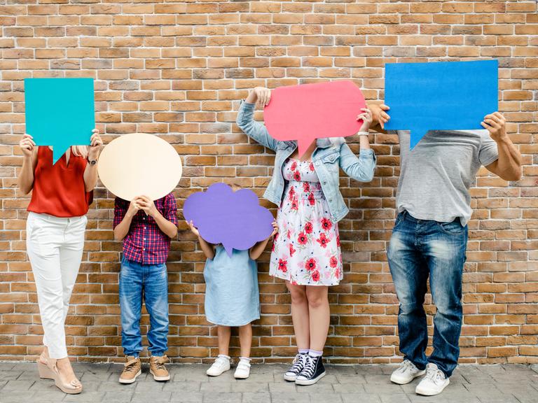 Jede Person der abgebildeten fünfköpfigen Familie hält eine Sprechblase aus farbiger Pappe vor dem Gesicht. Die Gesichter sind dabei nicht zu erkennen, es scheinen drei Kinder und zwei Erwachsene zu sein.