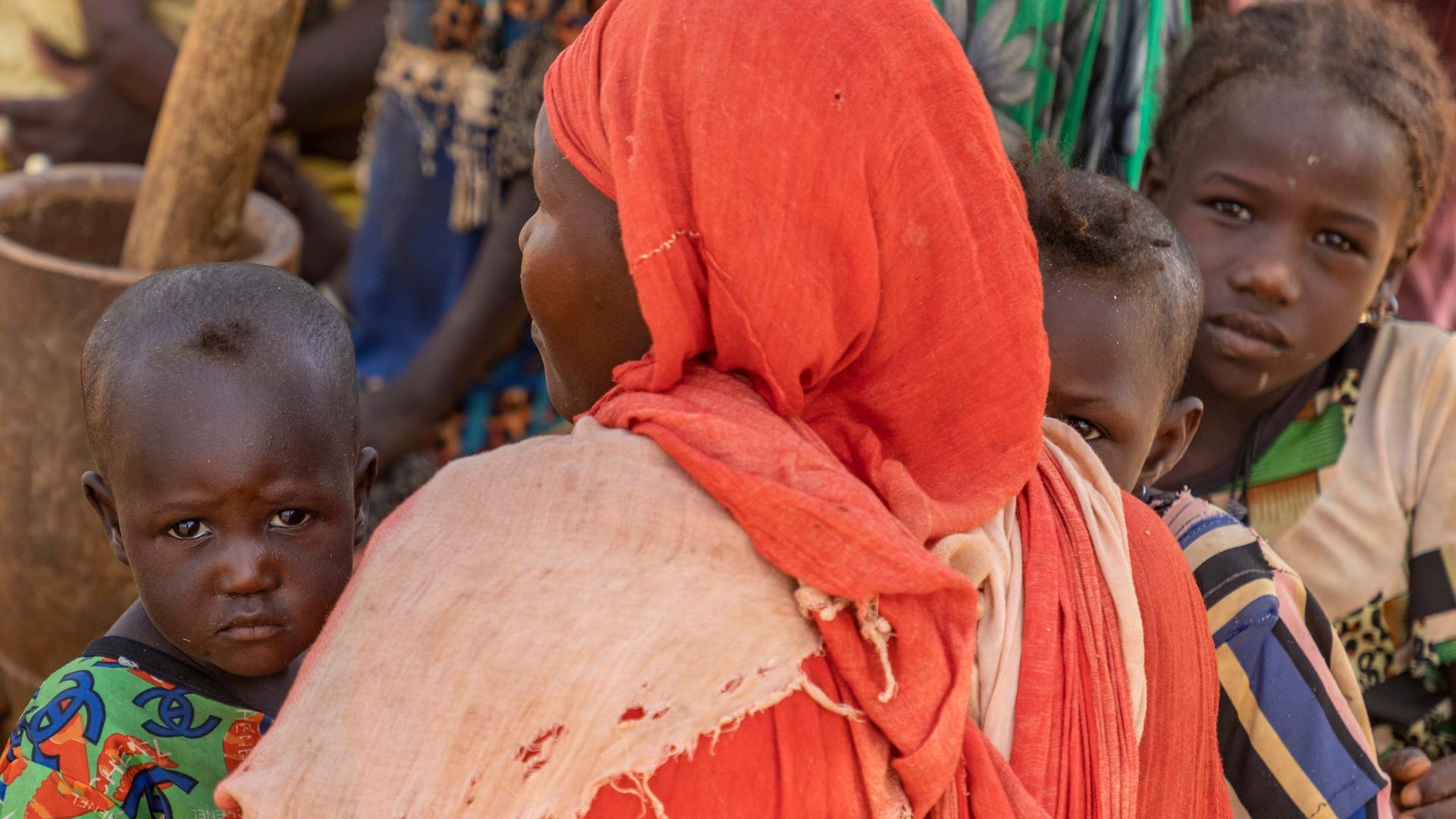 Eine Frau in Niger mit orangem Kopftuch hält ein Baby im Arm, hinter ihr zwei weitere Kinder. 