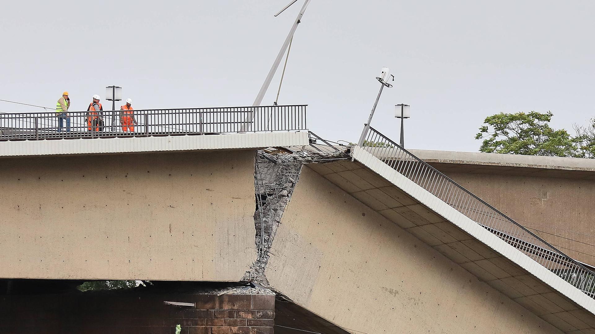 Blick auf die eingestürzte Carolabrücke in Dresden, Sachsen. Zu sehen ist der Beton auf einem Träger. Auf der einen Seite stehen drei Personen, vermutlich Männer in neonfarbener Kleidung.