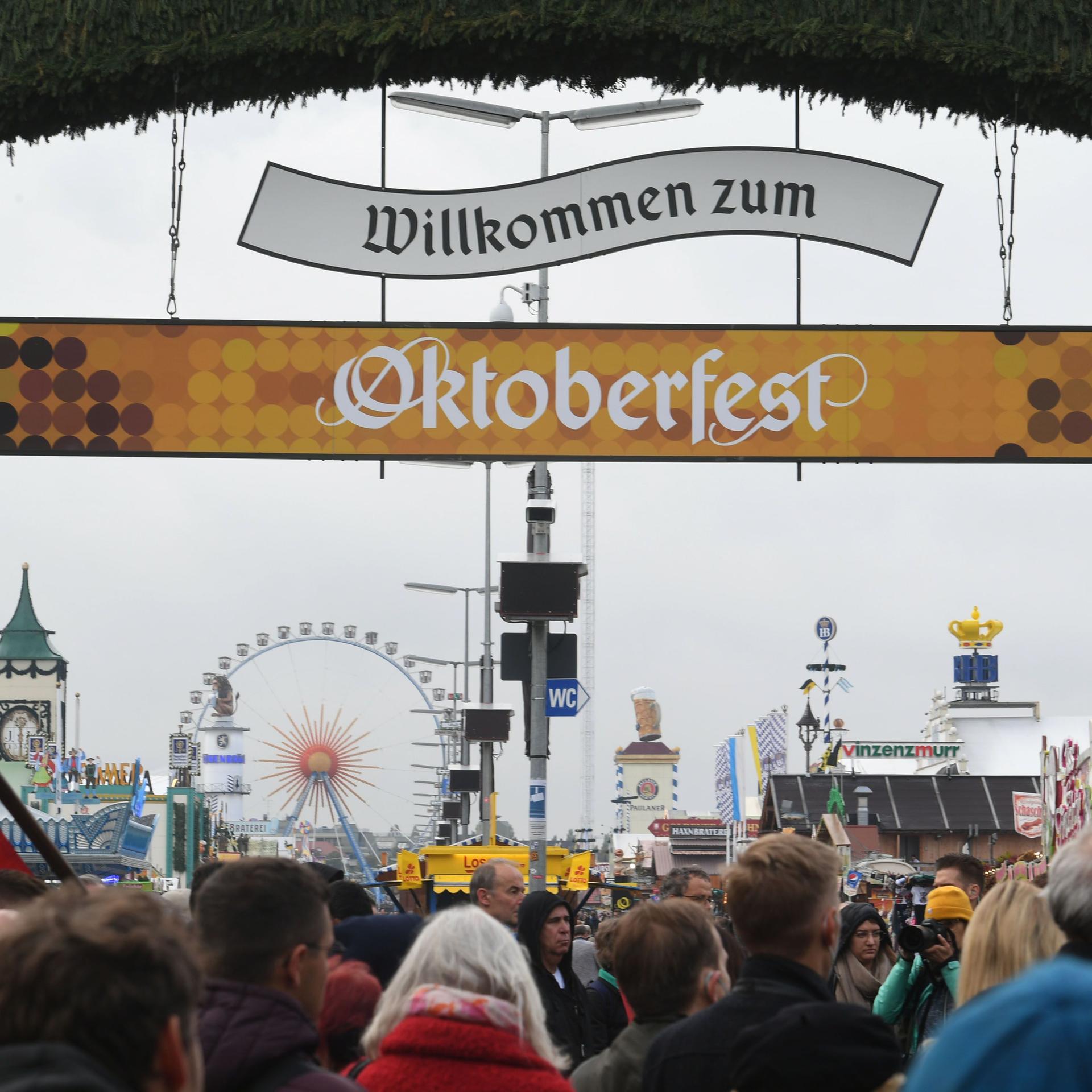 Über den hereinströmenden Besuchern hängen zwei Banner mit der Aufschrift "Willkommen zum Oktoberfest". Im Hintergrund ein Riesenrad und weitere Schausteller-Geschäfte.