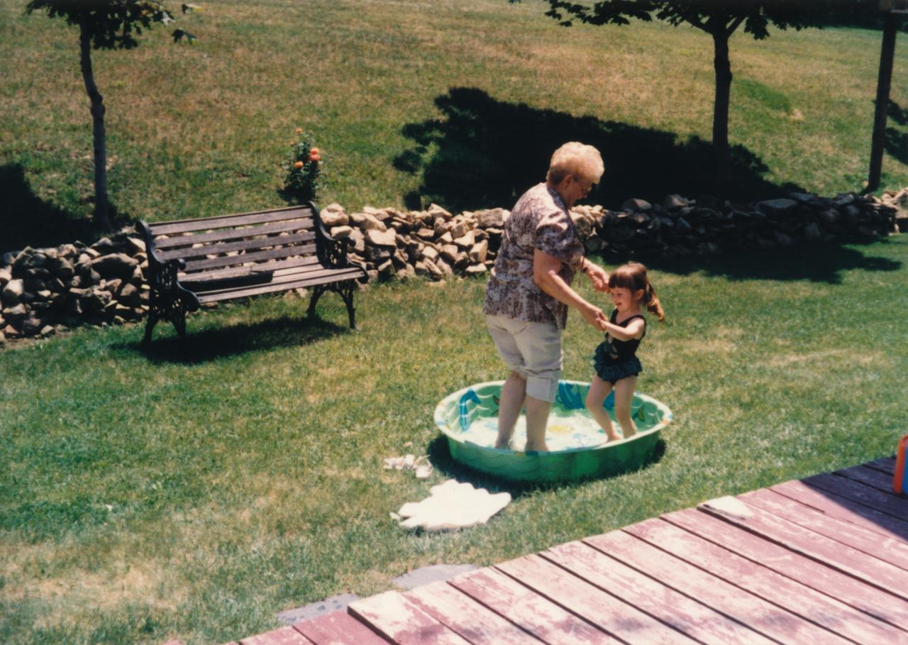 Eine Großmutter spielt mit Ihrer Enkelin draußen auf einer grünen Wiese in einem Planschbecken. Das Foto hat eine Retro-Anmutung. Es wurde vermutlich in den 1970er-Jahren aufgenommen.
