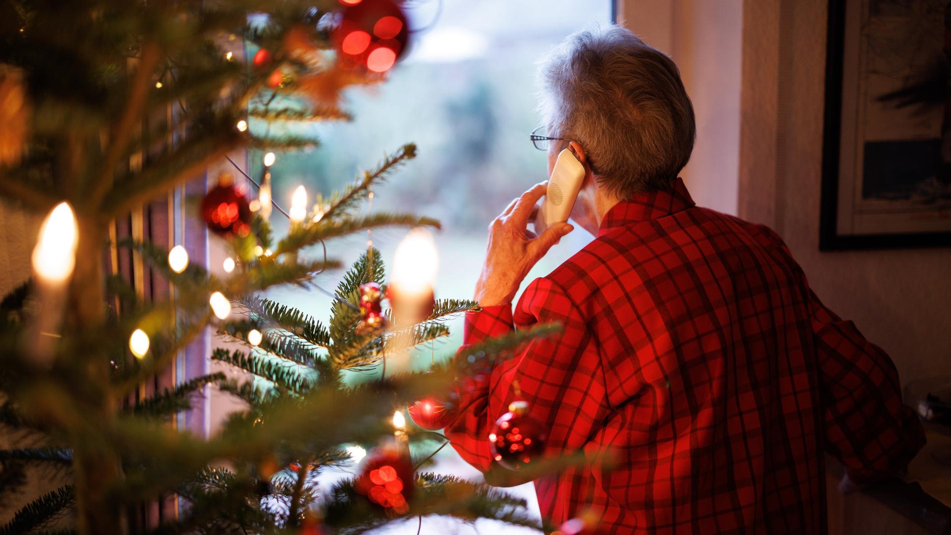 Eine ältere Dame steht mit einem Telefon am Ohr neben einem geschmückten Weihnachtsbaum und blickt aus einem Fenster der Wohnung.