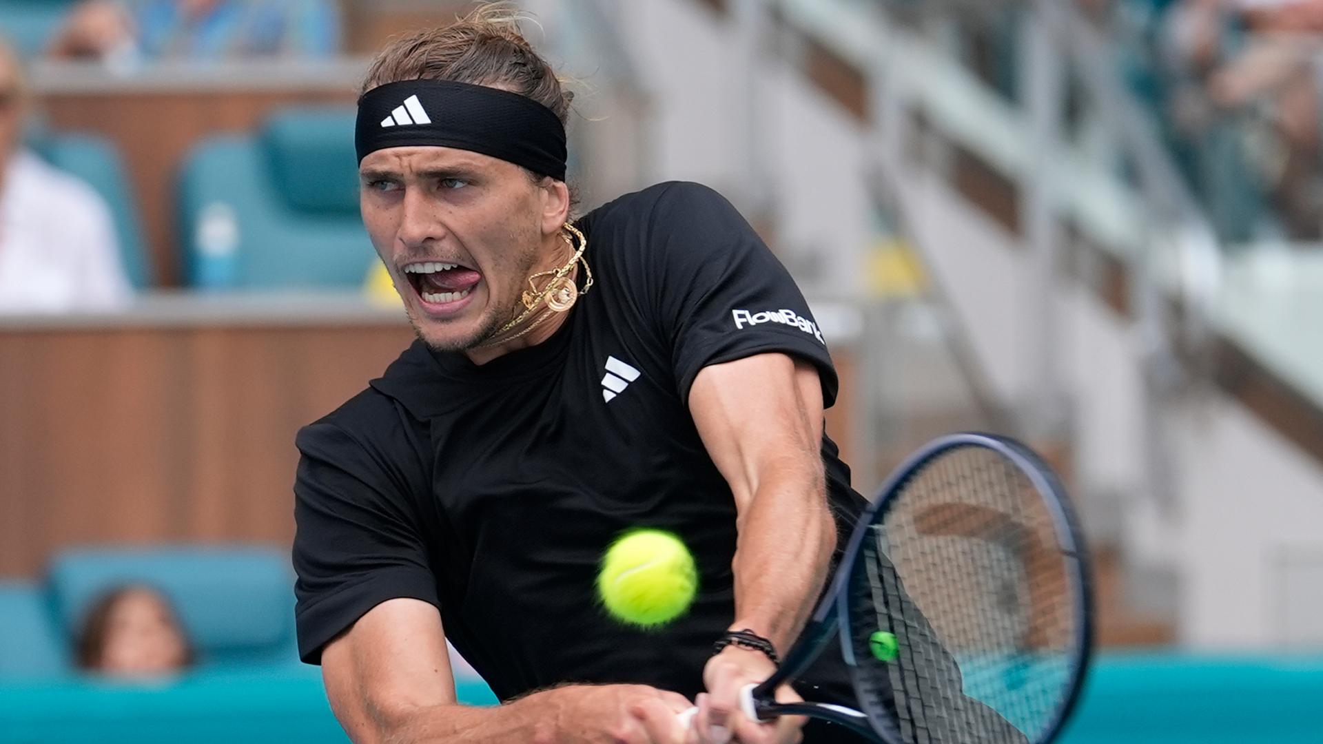 Alexander Zverev trägt ein Stirnband und Sportkleidung. Er schlägt den Tennisball mit seinem Schläger und verzieht dabei das Gesicht.