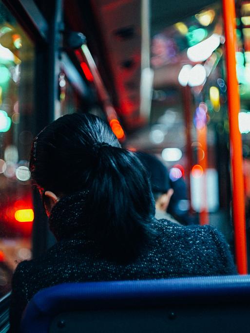 Rückansicht einer Frau, die in einem Bus sitzt - die farbige Beleuchtung der Straßen spiegelt sich in den Fenstern.