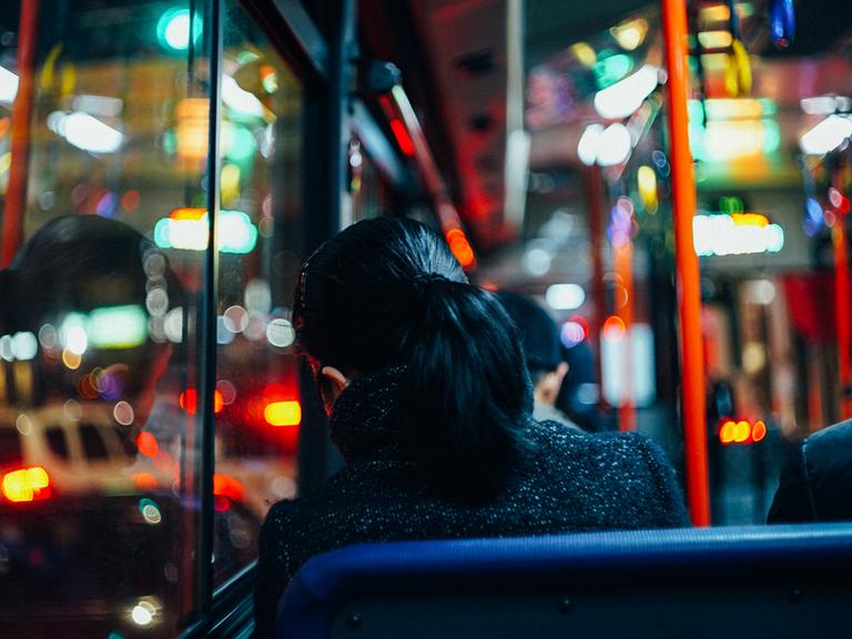 Rückansicht einer Frau, die in einem Bus sitzt - die farbige Beleuchtung der Straßen spiegelt sich in den Fenstern.