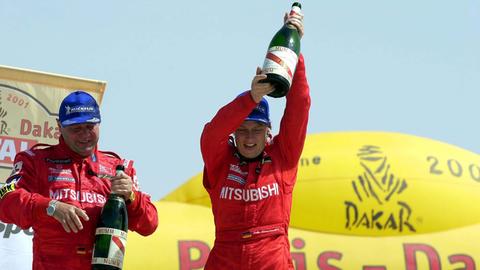 Jutta Kleinschmidt steht rechts neben ihrem Beifahrer Andreas Schulz. Beide halten Magnumflaschen Champagner in den Händen.