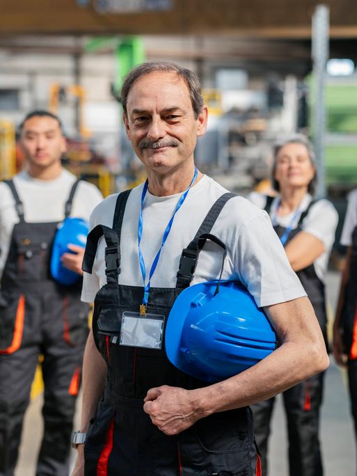 Ein lächelnder Industriearbeiter der Generation Boomer steht im Blaumann und mit gelbem Helm vor einer Gruppe von jüngeren Kolleginnen.