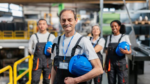 Ein lächelnder Industriearbeiter der Generation Boomer steht im Blaumann und mit gelbem Helm vor einer Gruppe von jüngeren Kolleginnen.