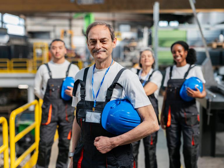 Ein lächelnder Industriearbeiter der Generation Boomer steht im Blaumann und mit gelbem Helm vor einer Gruppe von jüngeren Kolleginnen.