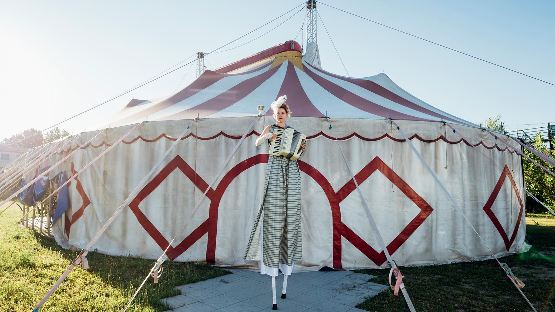 Ein Zirkus-Zelt steht auf einer Wiese. Davor steht eine Frau. Sie steht auf Stelzen und hat ein Akkordeon in der Hand.