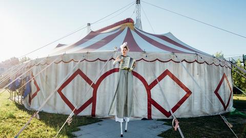 Zirkus bezaubert und ist atemberaubendes Spiel. Wer sind die Menschen, die hier arbeiten? Zu sehen: Eine Frau auf Stelzen steht vor einem Zirkuszelt und spielt Akkordeon. 