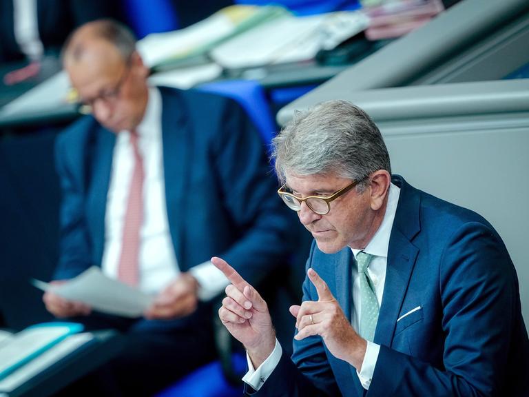 Wolfgang Weimer, Kulturstaatsminister, spricht in der Generaldebatte des Bundestags. Er hält die beiden Zeigefinger vor sich. Im Hintergrund ist verschwommen Friedrich Merz.