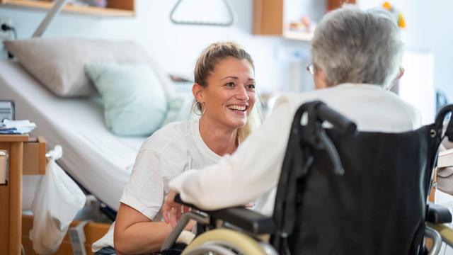 Eine junge Pflegerin lacht eine alte Frau im Roll-Stuhl an.